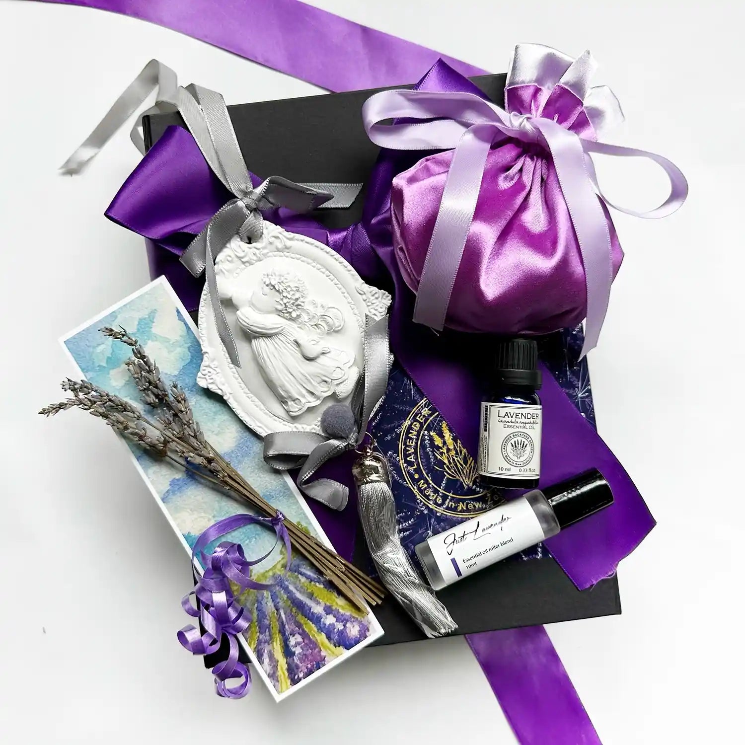 Gift set wrapped in a black gift box with a purple ribbon, featuring Just Lavender Essential Oil Rollerball, Aromatherapy Pure Lavender Essential Oils, Angel Scented Stone Diffuser, Luxury Satin Lavender Sachet Bag, Mini Dried Lavender Flowers, and Lavender Field Bookmark.