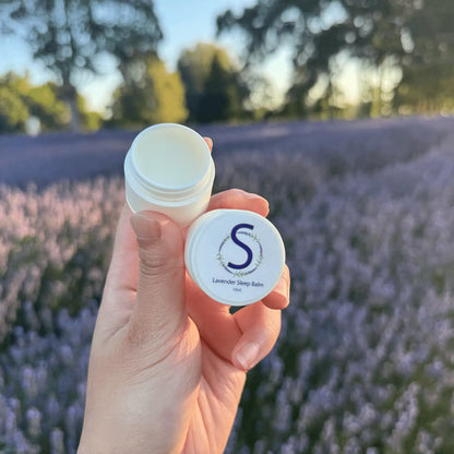 Lavender Sleep Balm with open lid showcasing inner texture, held by hand against a lavender field at sunset at Lavender Backyard Garden, NZ.