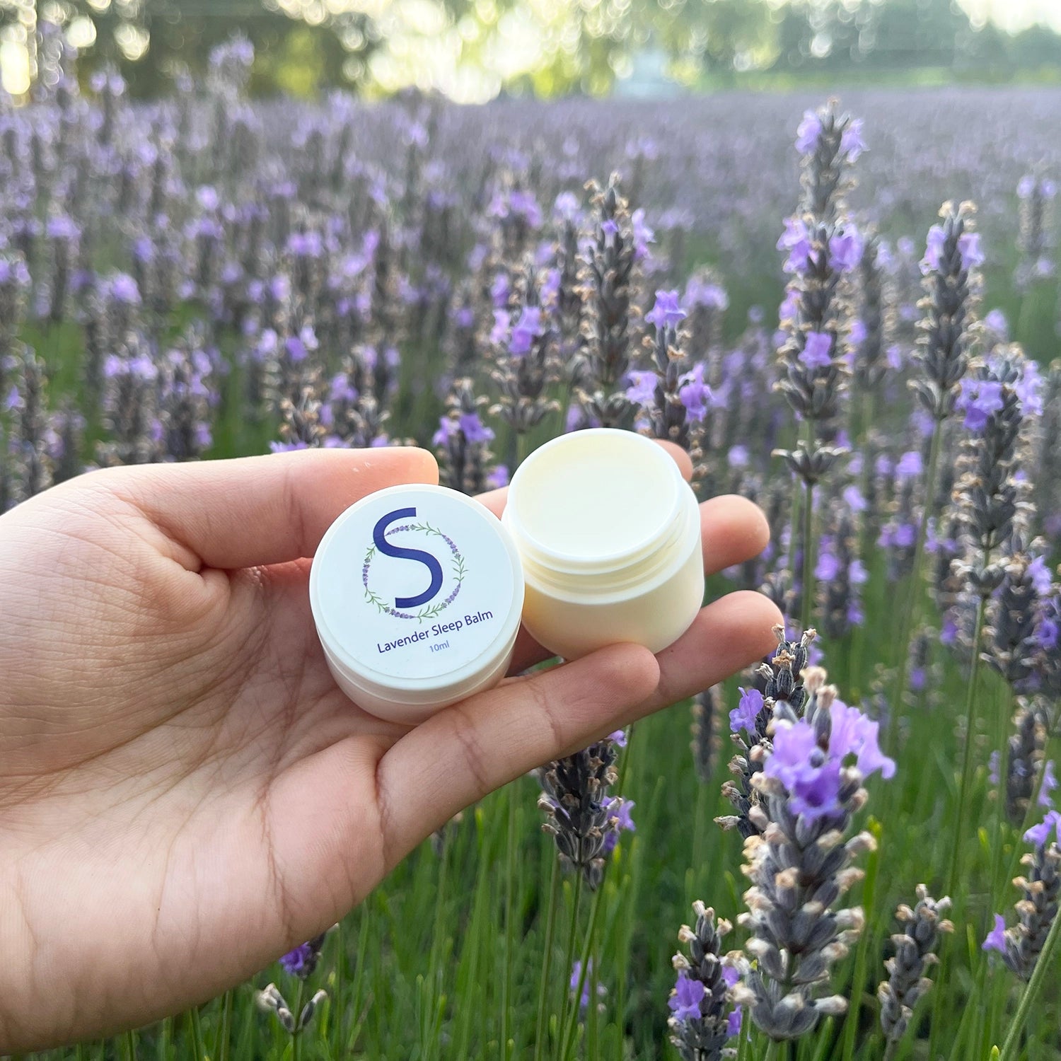 Open lid revealing the texture of Lavender Sleep Balm scented by essential oils, set against a backdrop of a purple lavender field at Lavender Backyard Garden.