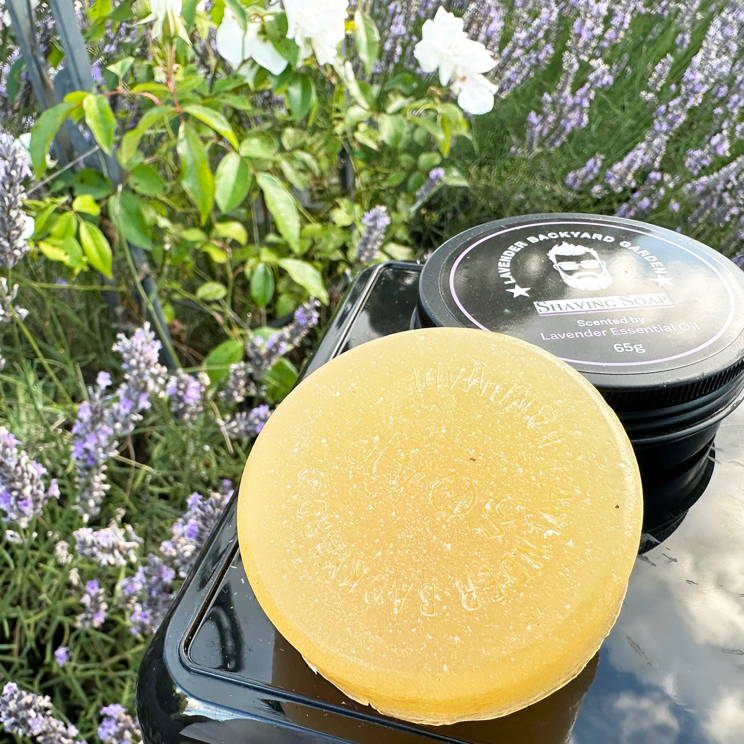 Lavender Soothing Shaving Soap with Shea Butter, placed against lavender blooms and white roses.