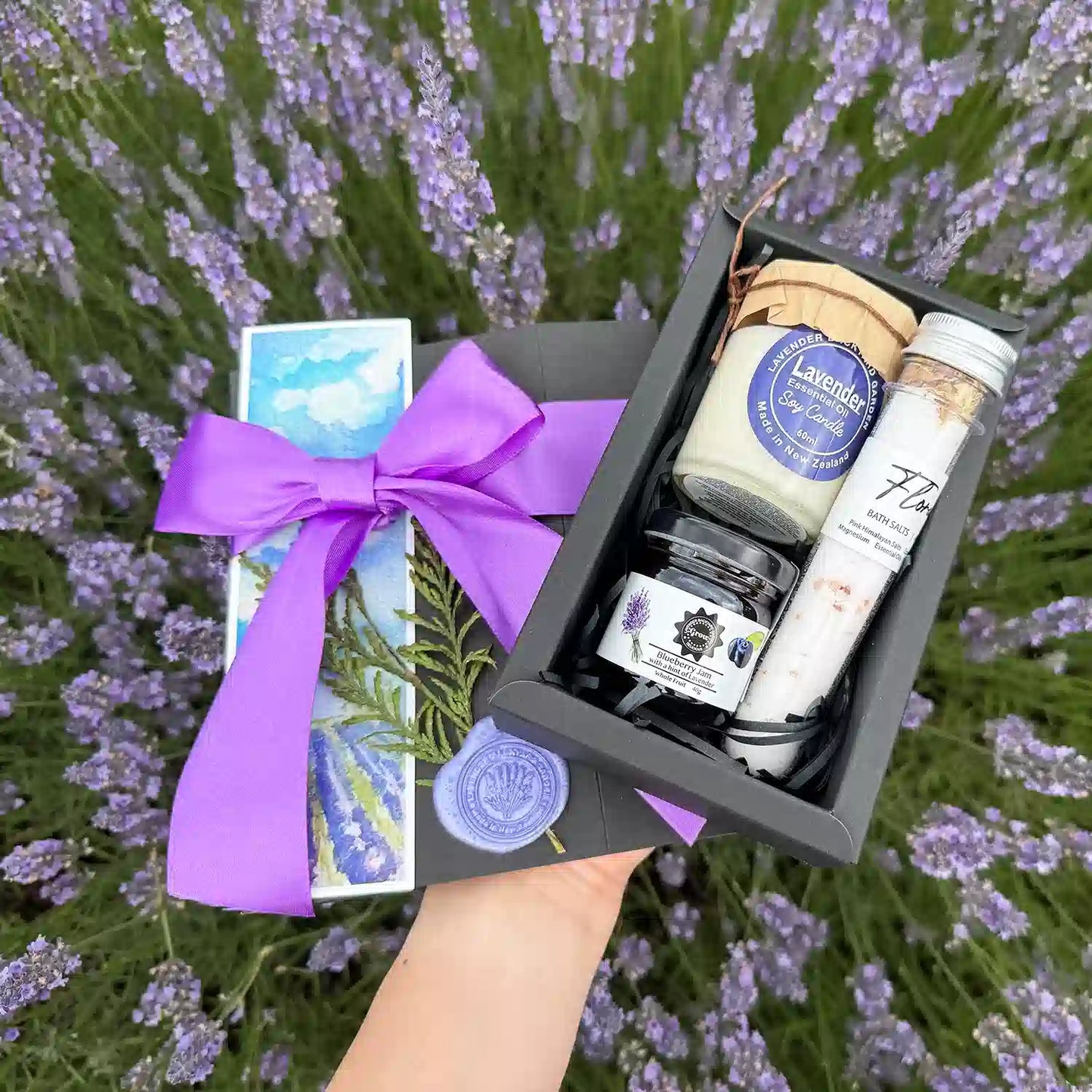 Lavender Sweet N Divine Gift Box - Small Gift featuring Floral Bath Salts Tube, Blueberry Jam with a hint of Lavender, Lavender Essential Oil Candle 60ml, and Lavender Field Bookmark against blooming lavender bushes.