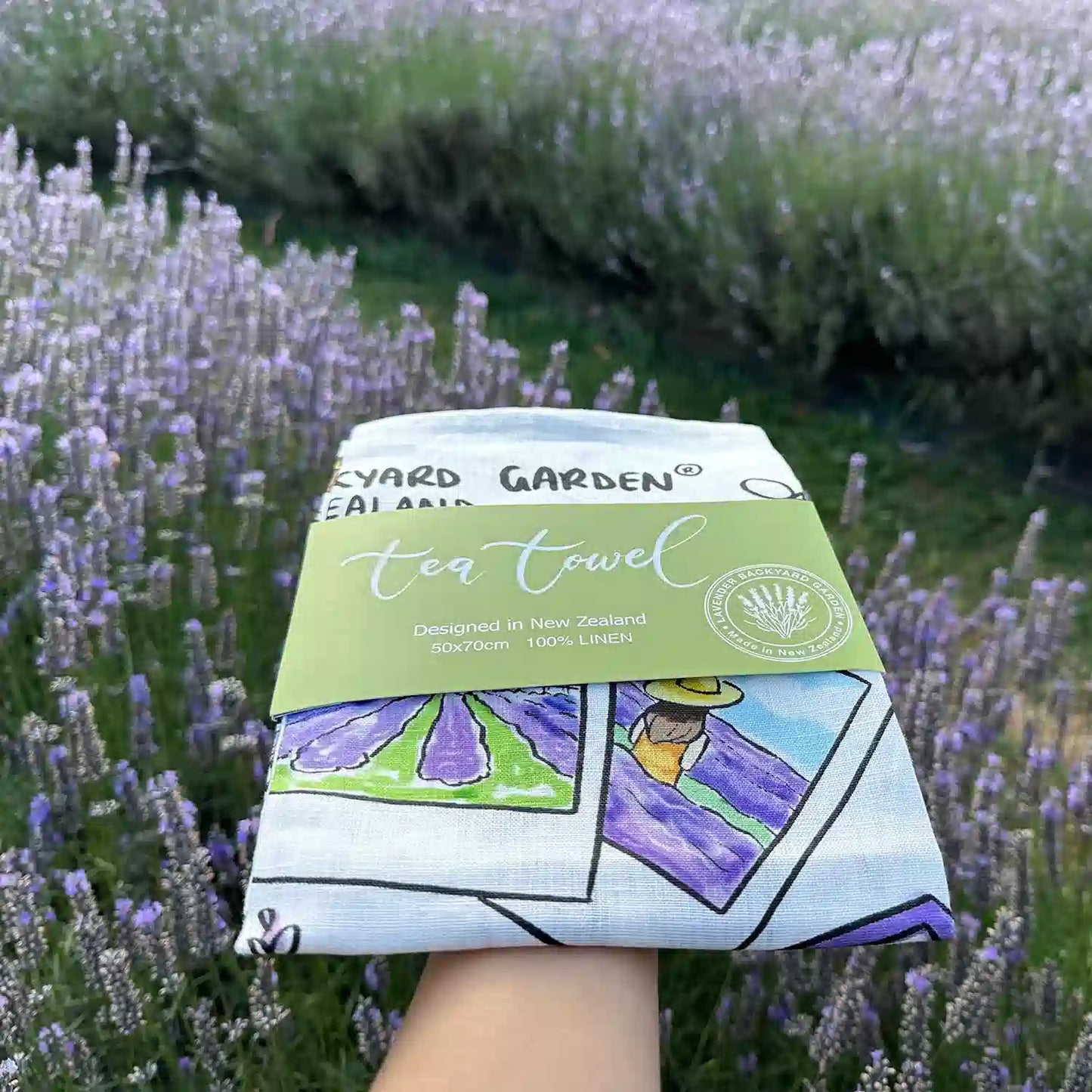 A flat Linen Tea Towel - Memory of Lavender Farm with a green package banner, held by hand against a backdrop of purple lavender fields at Lavender Backyard Garden, NZ close up view.