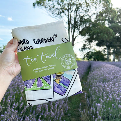 Close-up view of the folded Linen Tea Towel - Memory of Lavender Farm with a green package banner, placed against a lavender field backdrop at Lavender Backyard Garden, NZ.