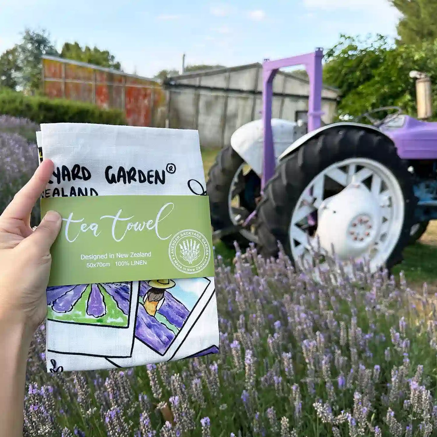 Folded Linen Tea Towel - Memory of Lavender Farm with a green package banner against purple tractors, lavender bloomings, and the farm shed at Lavender Backyard Garden, NZ.