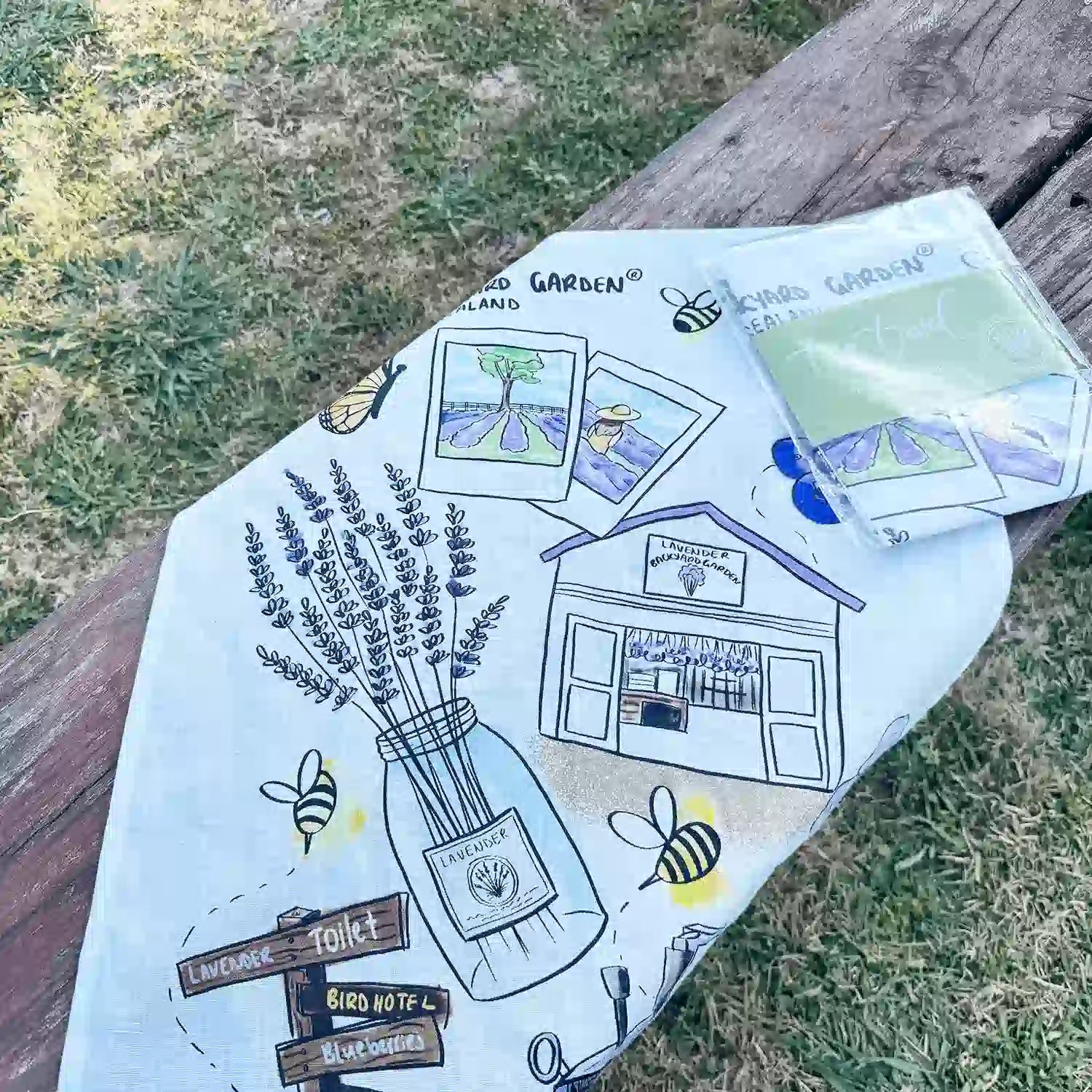 Two linen tea towels, one unfolded and the other in its package, placed on a wooden bench with a backdrop of lavender field at Lavender Backyard Garden.