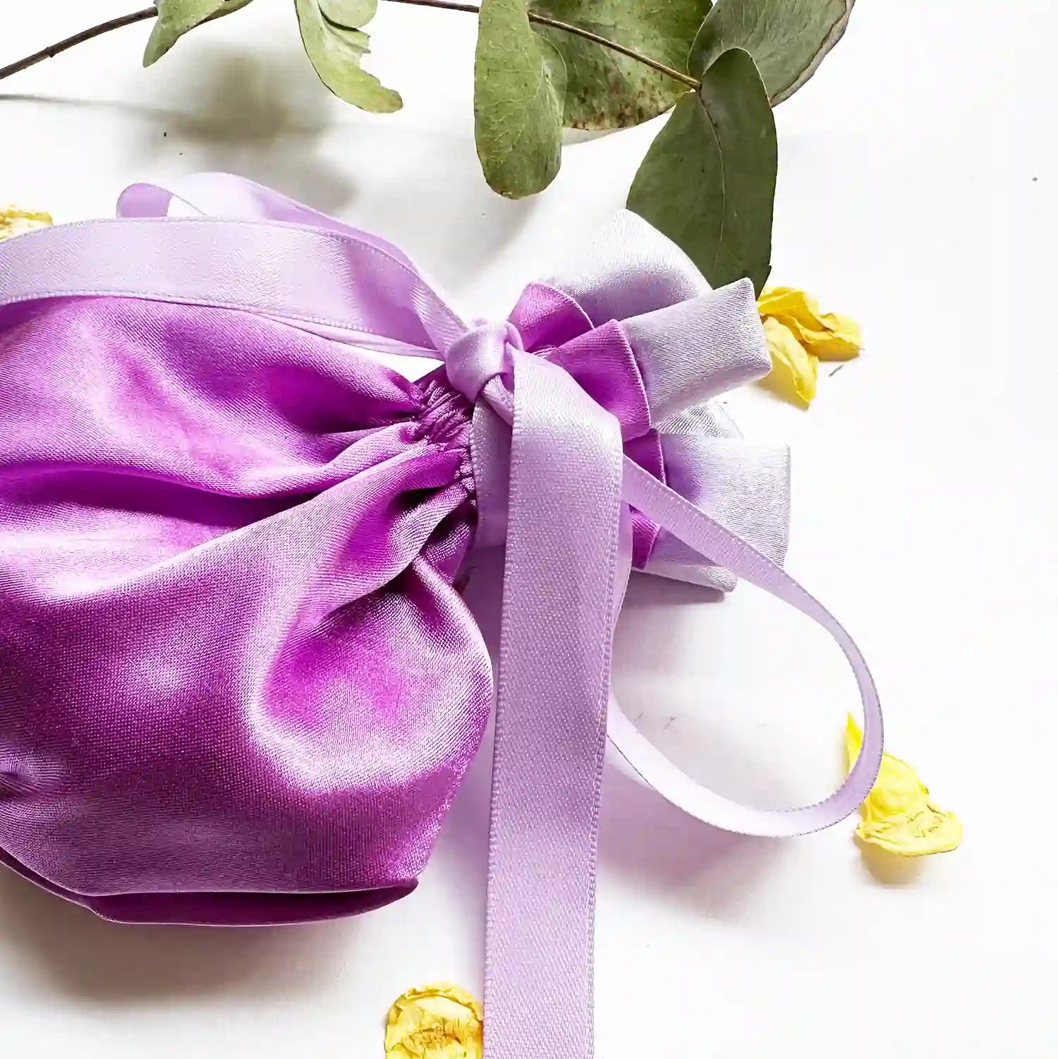 Close-up view of a satin lavender sachet bag with a decorative background of eucalyptus leaves and rose petals.