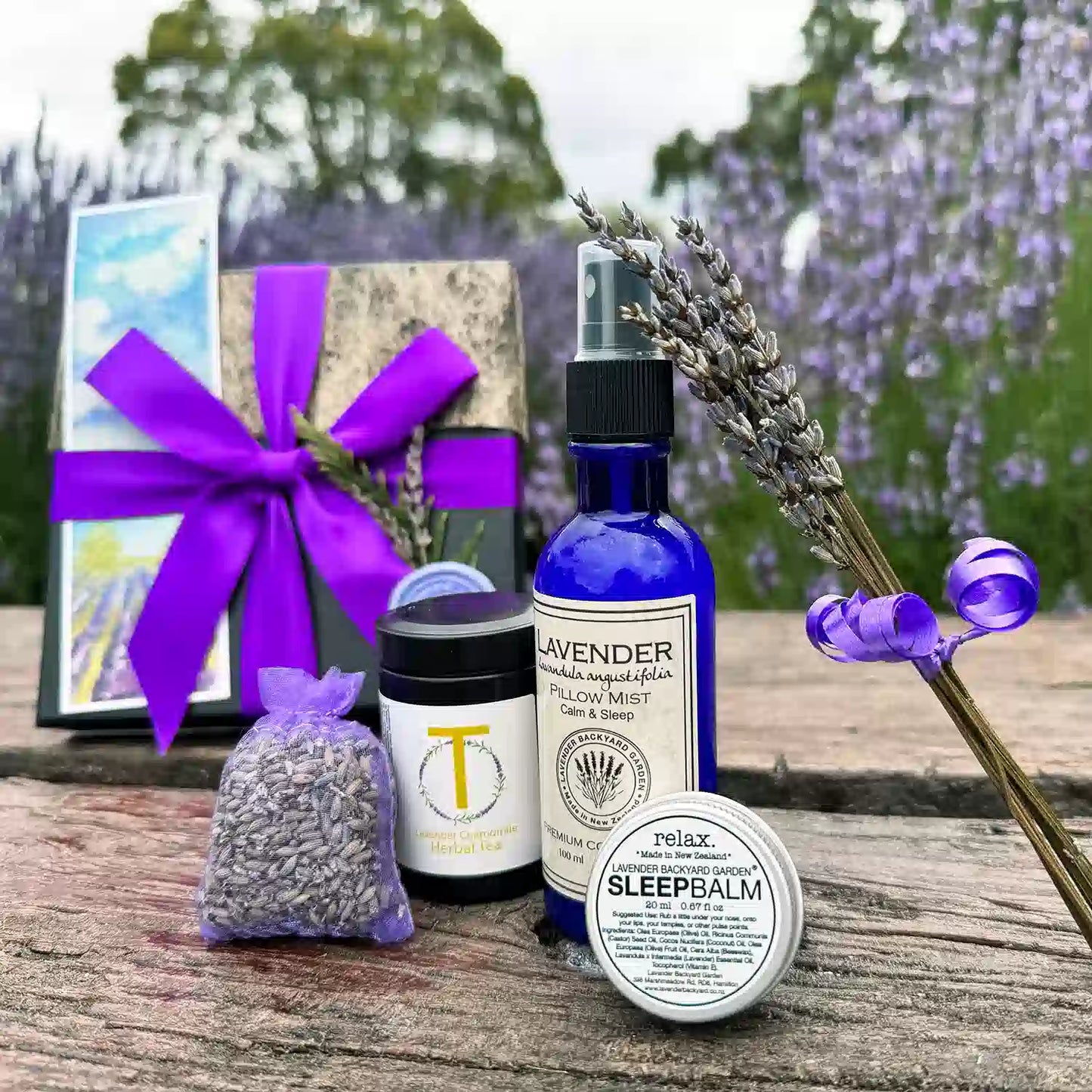 Lovely Lavender Therapy Giftset for Women scented by essential oils, placed on a wooden bench featuring products "Dried Lavender Sachet, Lavender Sleep Balm, Lavender Pillow Mist, Lavender & Chamomile Herbal Tea - Loose Leaf (Travel), Mini Dried Lavender Flowers, Lavender Field Bookmark" against a purple blooming lavender field.