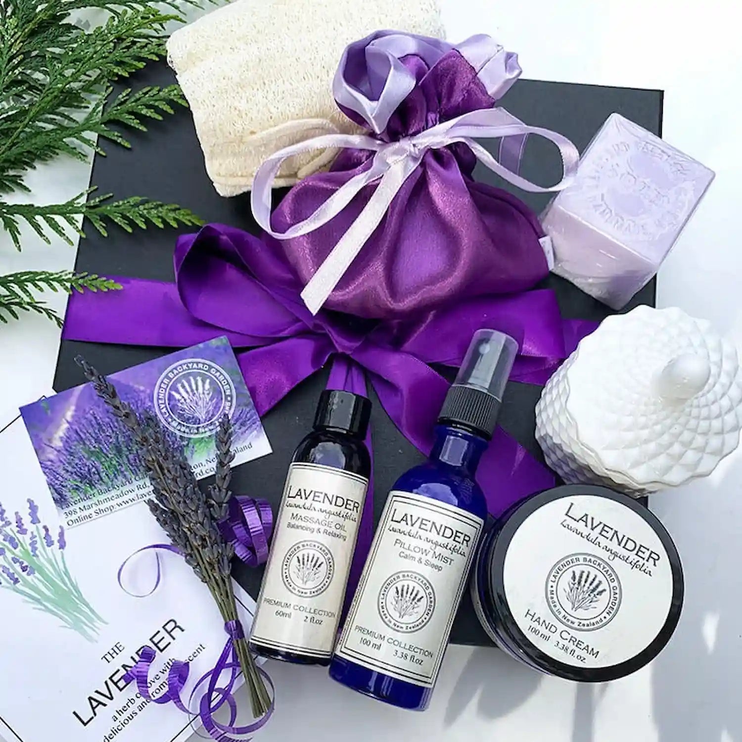 Luxuries Lavender Gift Set, featuring essential oil-scented products including lavender sleep spray, hand cream, massage oil, coconut wax candle, lavender goat milk soap, dried flower satin sachet bag, and a greeting card, all arranged against a background of a purple ribbon gift box.