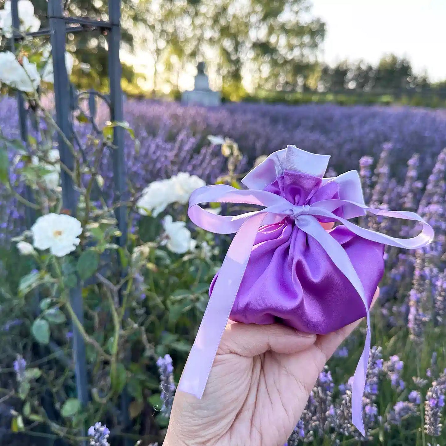 Luxury Purple Satin Silk Lavender Sachet Bag placed beside white roses with a lavender field at sunset in Lavender Backyard Garden, NZ.