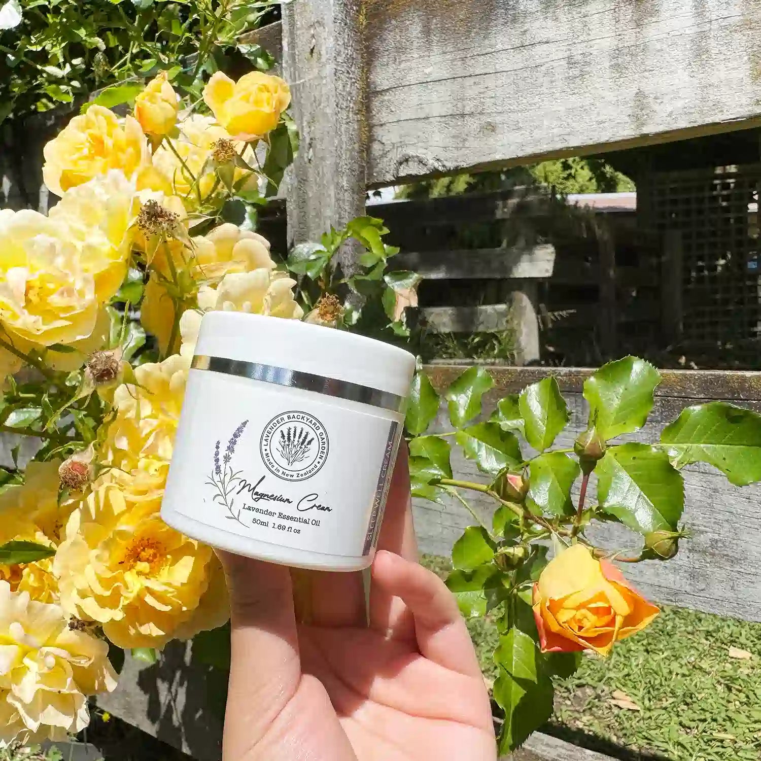 Magnesium Sleep Cream, scented by lavender essential oil, held by hand in front of a farm fence with yellow roses.