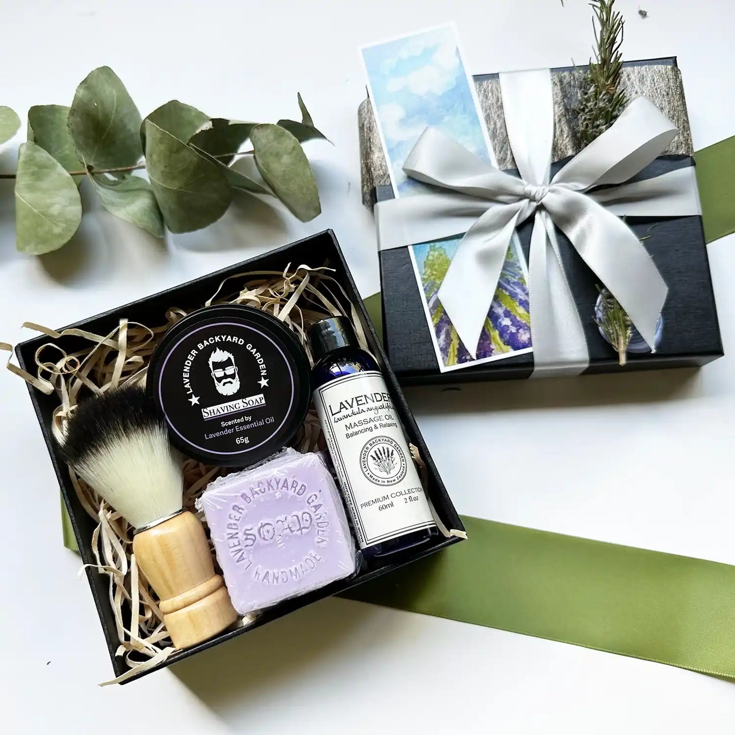 A stylish Man Therapy Lavender Gift Box featuring lavender shaving soap for men, lavender handmade soap, a shaving brush, and massage oil, all elegantly wrapped in a black gift box with a silver ribbon, accompanied by a lavender field bookmark.