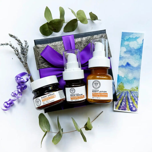 A beautifully arranged gift set featuring Manuka honey body lotion, lavender-scented facial cream, and neck serum, all elegantly wrapped with a purple ribbon and accompanied by a lavender field bookmark.