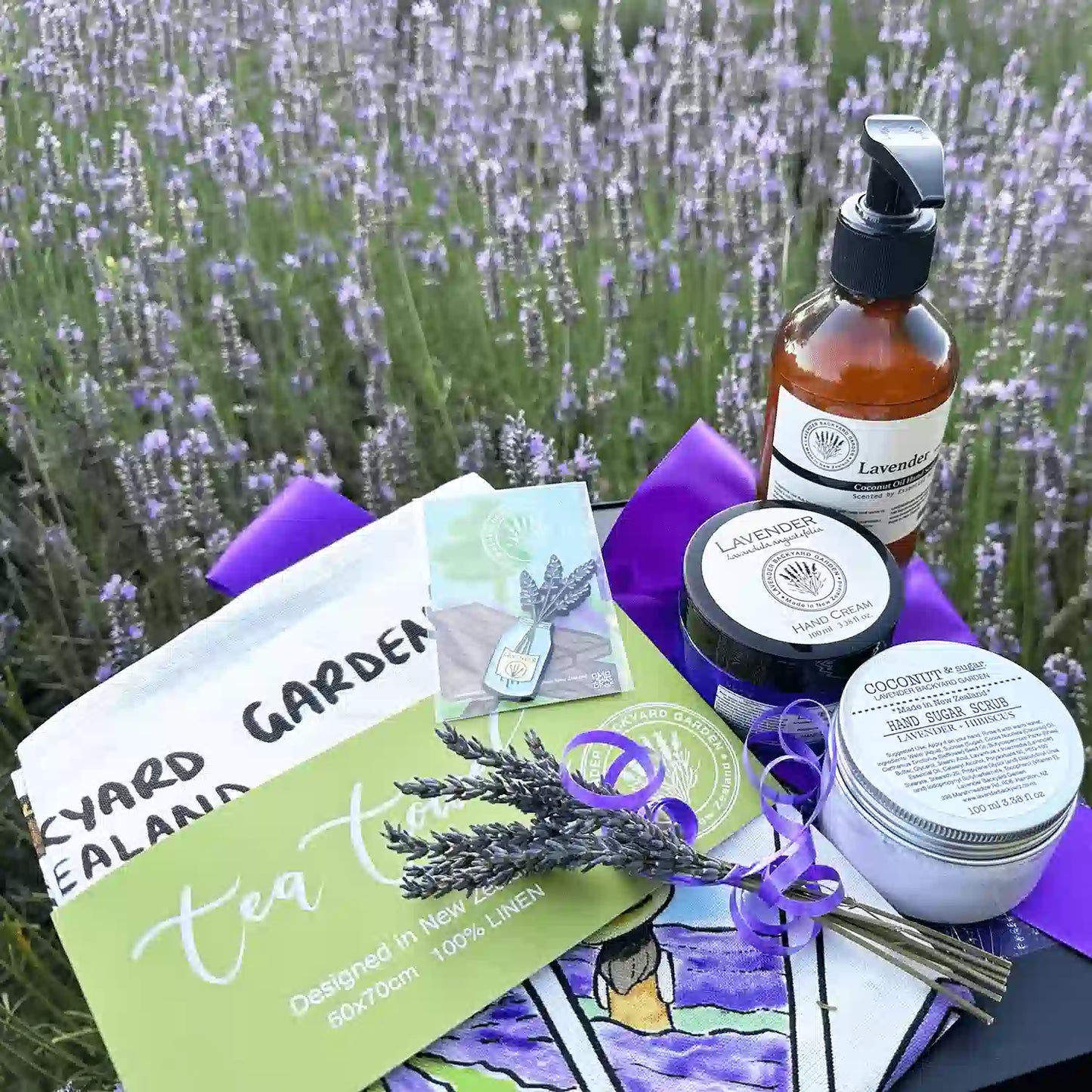 Mother's Kitchen Comforting Treat Lavender Gift Sets placed against purple lavender bloomings, featuring products: Lavender Sugar Scrub, Lavender Hand Cream for Dry Hands, Tea Towel - Memory of Lavender Farm, Therapy Lavender Hand Wash, A Jar of Lavender Fridge Magnets, Mini Lavender Dry Flower, and Lavender Field Bookmark.