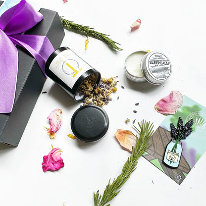 Close-up view of a "Nice Thoughtful Gift Set for Friends," featuring a black gift box with a purple ribbon. The set includes Lavender & Camomile Herbal Tea (Mini), Lavender Sleep Balm, and a Jar of Lavender Fridge Magnets, surrounded by rose petals and rosemary as background decor.