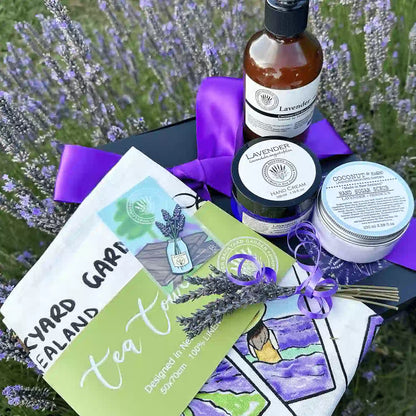 Close-up view of the Nurturing Kitchen Treats for Mother Gift Idea, featuring products Lavender Sugar Scrub, Lavender Hand Cream for Dry Hands, Tea Towel - Memory of Lavender Farm, Therapy Lavender Hand Wash, A Jar of Lavender Fridge Magnets, Mini Lavender Dry Flower, and Lavender Field Bookmark, beautifully arranged in a purple ribbon gift box against a backdrop of blooming lavender fields.