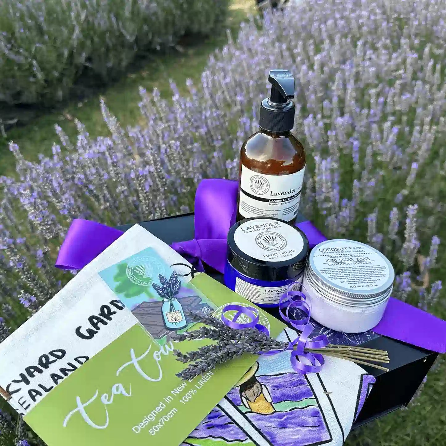 Nurturing Kitchen Treats for Mother Gift Idea featuring Lavender Sugar Scrub, Lavender Hand Cream for Dry Hands, Tea Towel - Memory of Lavender Farm, Therapy Lavender Hand Wash, A Jar of Lavender Fridge Magnets, Mini Lavender Dry Flower, and Lavender Field Bookmark, beautifully placed on a purple ribbon gift box against a blooming purple lavender field.