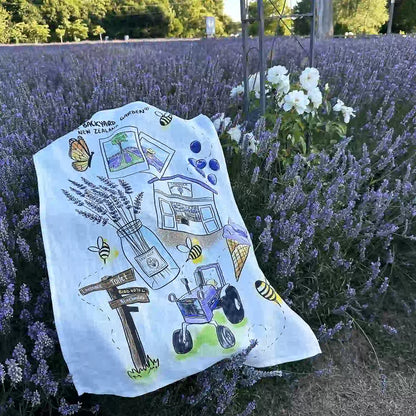 NZ-designed linen tea towel for Lavender Backyard Garden, draped over blooming lavender bushes with white roses in the foreground.