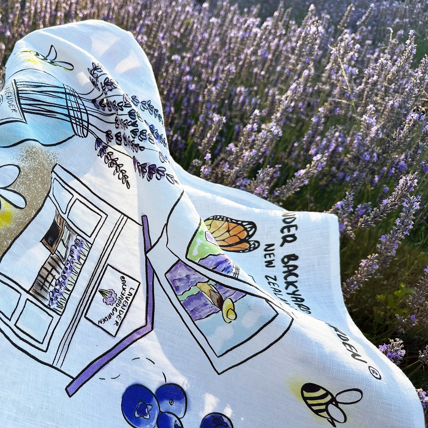 Close-up view of NZ farm design linen tea towel featuring "Memory of Lavender Farm," with detailed embroidery and textures, against a backdrop of blooming lavender at Lavender Backyard Garden, NZ.