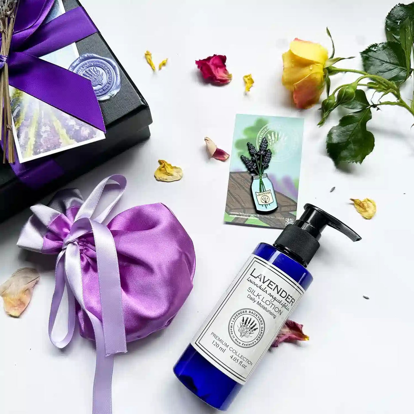 Close-up view of the Pamper & Relax Aroma Giftset in a black gift box with a purple ribbon, featuring moisturizing lavender body lotion, a luxury satin lavender sachet bag, and a jar of lavender fridge magnets. Background decor includes fresh yellow rose and rose petals.