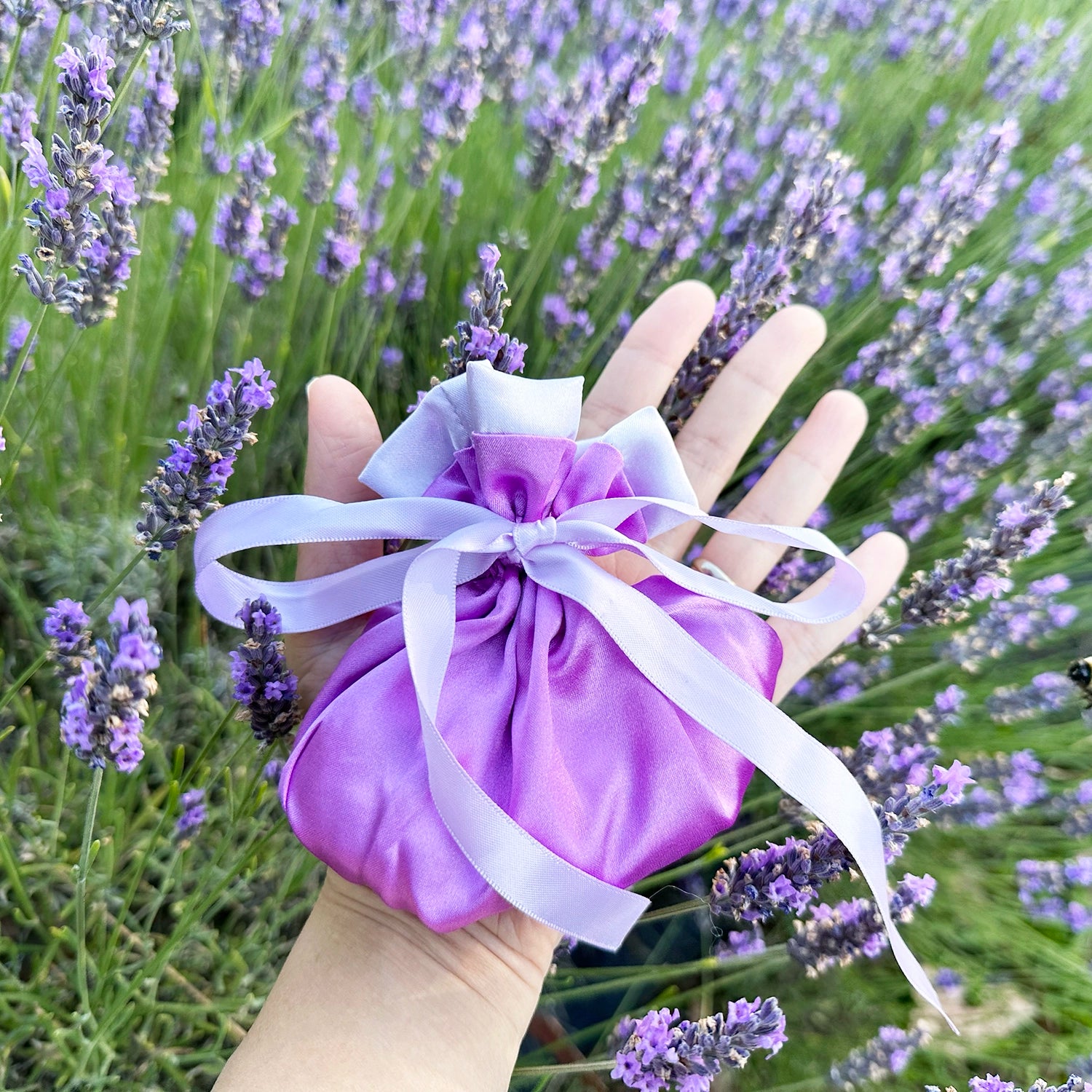 Purple satin scented sachet bag held in hand above blooming lavender flowers.