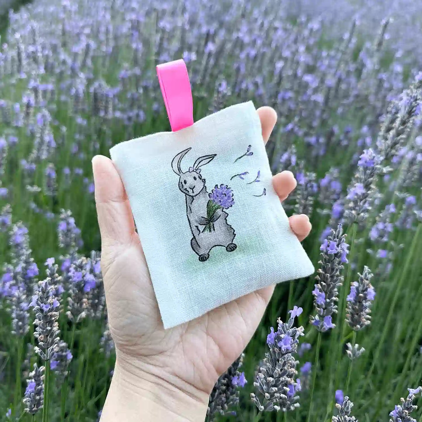 Rabbit Scented hanging sachets for closet and wardrobe, holding by hand against a backdrop of blooming lavender flowers at Lavender Backyard Garden, NZ.