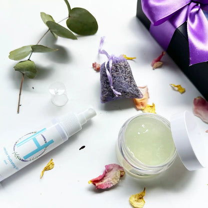 Close-up view of "Radiate & Restore Skin Care Gift Set" with an open lid showing the texture of Aloe Vera Face Massage Gel, alongside a Lavender Hydrosol Floral Water Face Toner (Travel), Dried Lavender Sachet, and Lavender Field Bookmark. The products are elegantly presented with a black gift box tied with a purple ribbon, with rose petals and eucalyptus leaves as background decor.