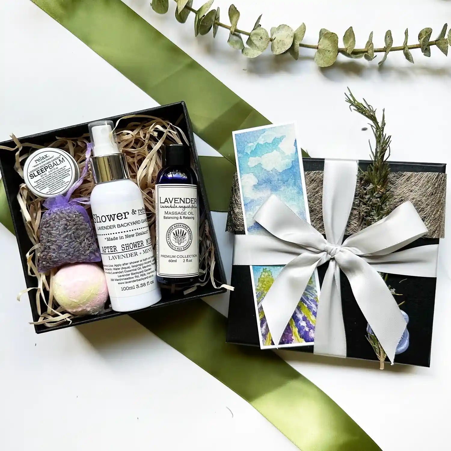 A beautifully arranged Relaxing Bath & Shower Gift Box featuring massage oil, after shower mist, and sleep balm, all scented with lavender essential oils. The box includes a lavender sachet and bath bomb, elegantly packed in a black gift box with a silver ribbon and a lavender field bookmark.