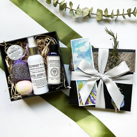 A beautifully arranged Relaxing Bath & Shower Gift Box featuring massage oil, after shower mist, and sleep balm, all scented with lavender essential oils. The box includes a lavender sachet and bath bomb, elegantly packed in a black gift box with a silver ribbon and a lavender field bookmark.