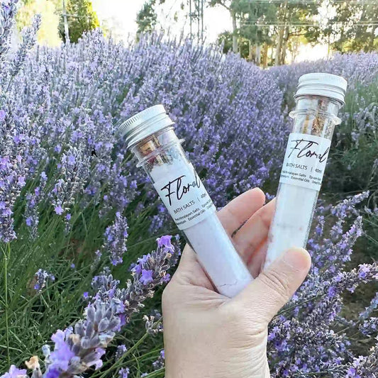 Two floral bath salt tubes, held by hand against a purple lavender blooming field at Lavender Backyard Garden, NZ.