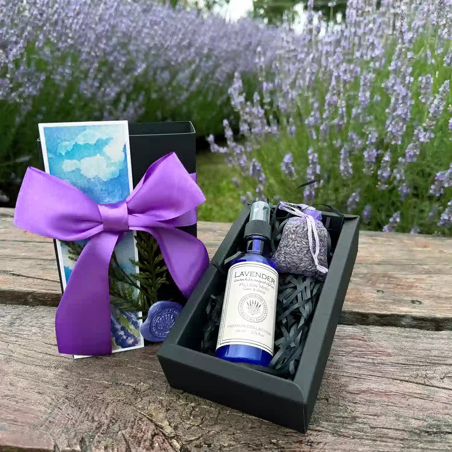 Restful Night Sleep Gift Box scented by lavender essential oils featuring Lavender Essential Oil Pillow Mist, Dried Lavender Sachet, and Lavender Field Bookmark, placed on a wooden bench against purple lavender blooming bushes.