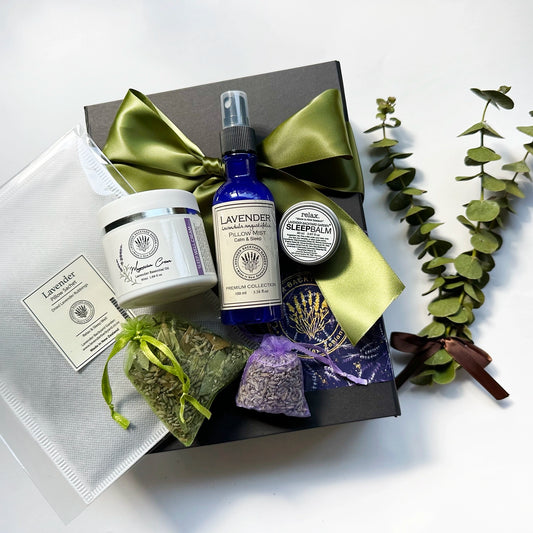 A beautifully arranged "Sleep Easy Comfort Gift Set" featuring magnesium oil face cream, sleep spray, sleep balm, lavender sachet, pillow sachet, and eucalyptus sachet, all elegantly wrapped in a black gift box with a green ribbon, accompanied by a lavender field bookmark.