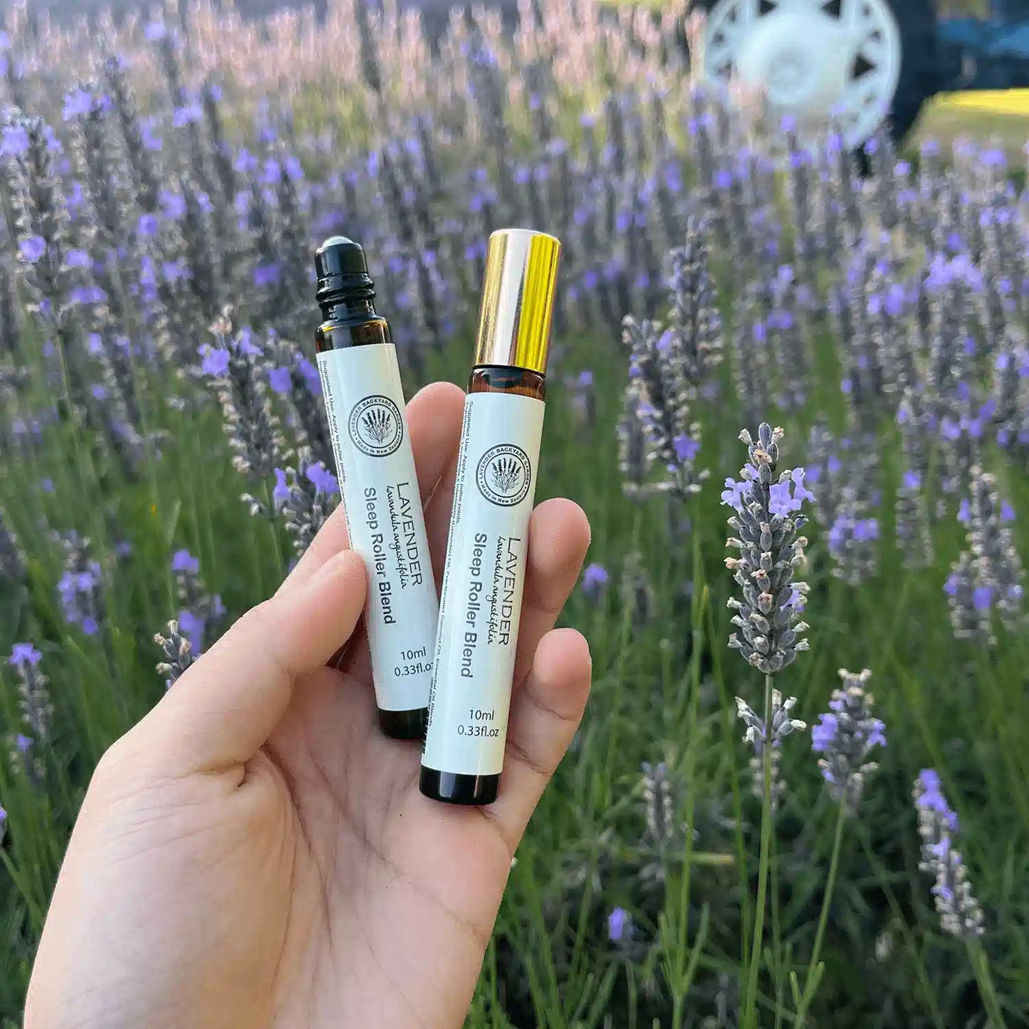 Two sleep roll-ons, one with the lid open showcasing the roller ball and the other closed, held by hand against a backdrop of blooming purple lavender fields at Lavender Backyard Garden, NZ.