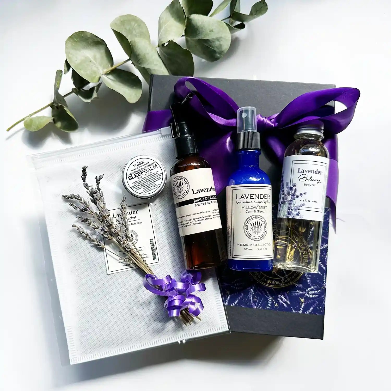 Sleep Support Essentials Gift Set presented in a black gift box wrapped with a purple ribbon, containing a lavender sleep sachet, lavender sleep spray, lavender bath oil, and lavender sleep balm, accompanied by mini dried lavender flowers and a lavender field bookmark.