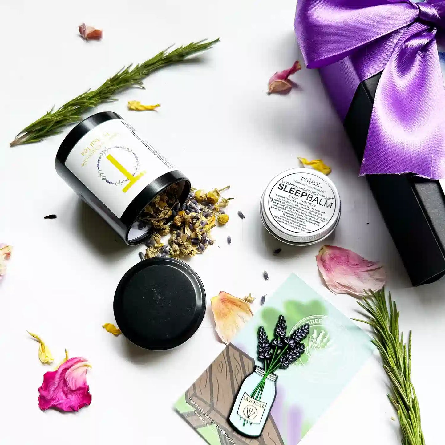 Close-up view of a "Small Thoughtful Lavender Gift" set, featuring a black gift box with a purple ribbon. The set includes Lavender & Camomile Herbal Tea (Mini), Lavender Sleep Balm, and a Jar of Lavender Fridge Magnets, surrounded by rose petals and rosemary as background decor.