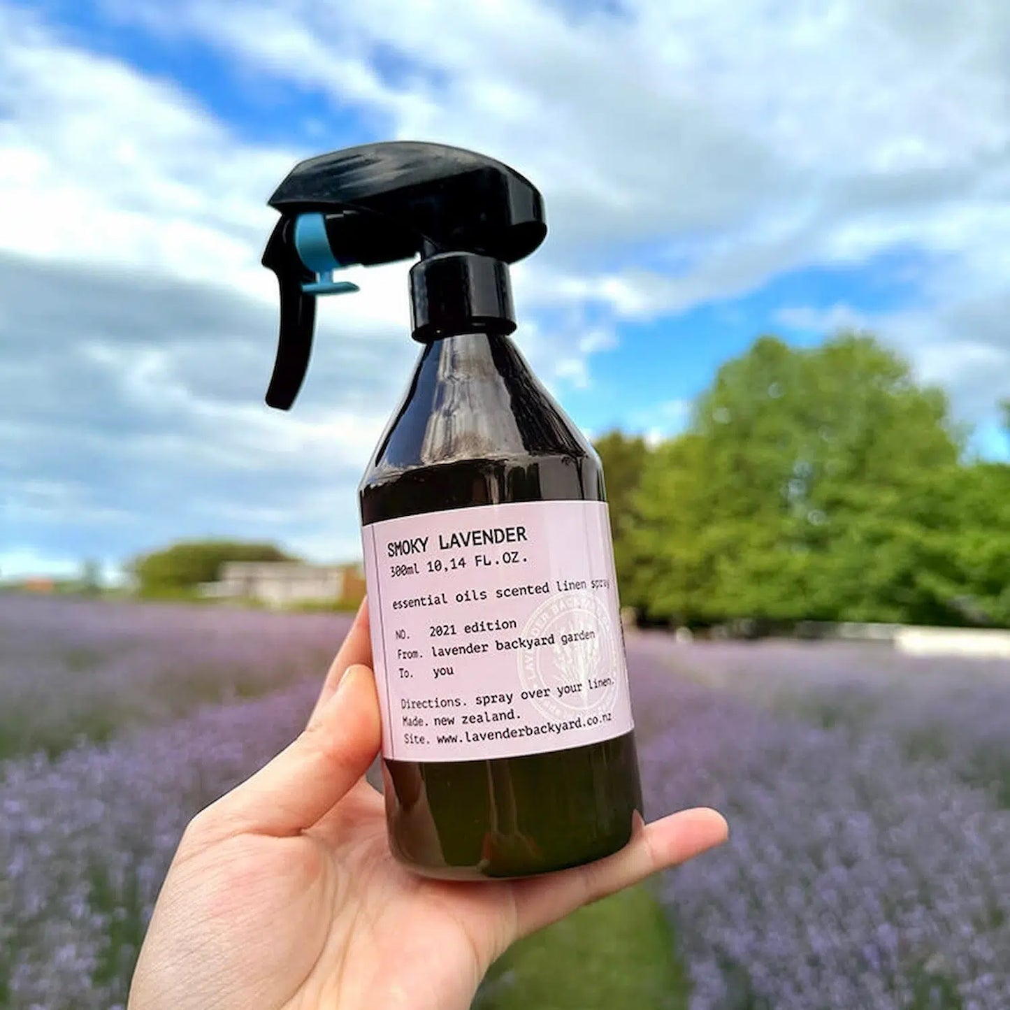 Smoky Lavender Linen Spray, scented by essential oils, displayed against a lavender field with a blue sky and clouds in the background.