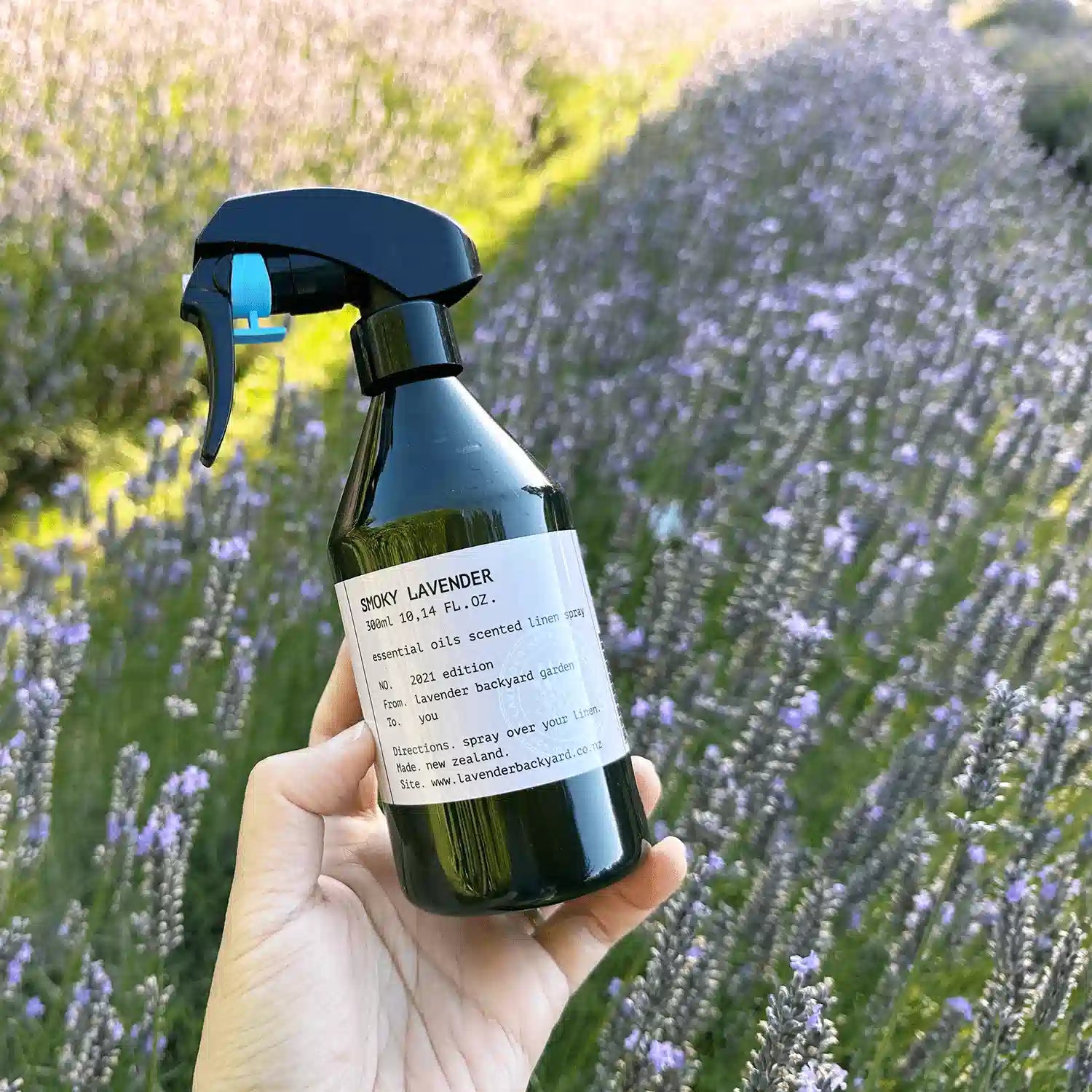 Smoky lavender natural linen spray bottle held by hand against a backdrop of blooming lavender flowers at Lavender Backyard Garden, NZ.