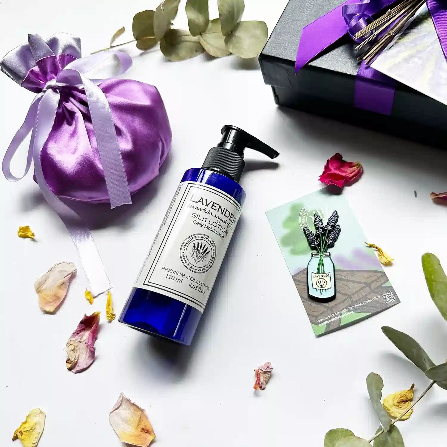  Close-up view of the Soothing Aroma Gift Hamper in a black gift box with a purple ribbon, featuring moisturizing lavender body lotion, a luxury satin lavender sachet bag, and a jar of lavender fridge magnets. Background decor includes eucalyptus leaves and rose petals.