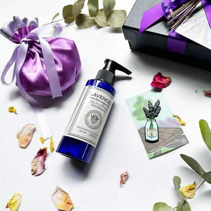  Close-up view of the Soothing Aroma Gift Hamper in a black gift box with a purple ribbon, featuring moisturizing lavender body lotion, a luxury satin lavender sachet bag, and a jar of lavender fridge magnets. Background decor includes eucalyptus leaves and rose petals.