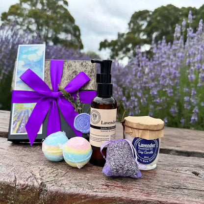 Supreme Lavender Bath Spa Gift Set placed on a wooden bench against blooming purple lavender, featuring Lavender Essential Oil Soy Candle, Lavender Essential Oil Bath Bomb, Luxury Lavender Bath Oils, Mini Lavender Dried Flowers, and Lavender Field Bookmark.