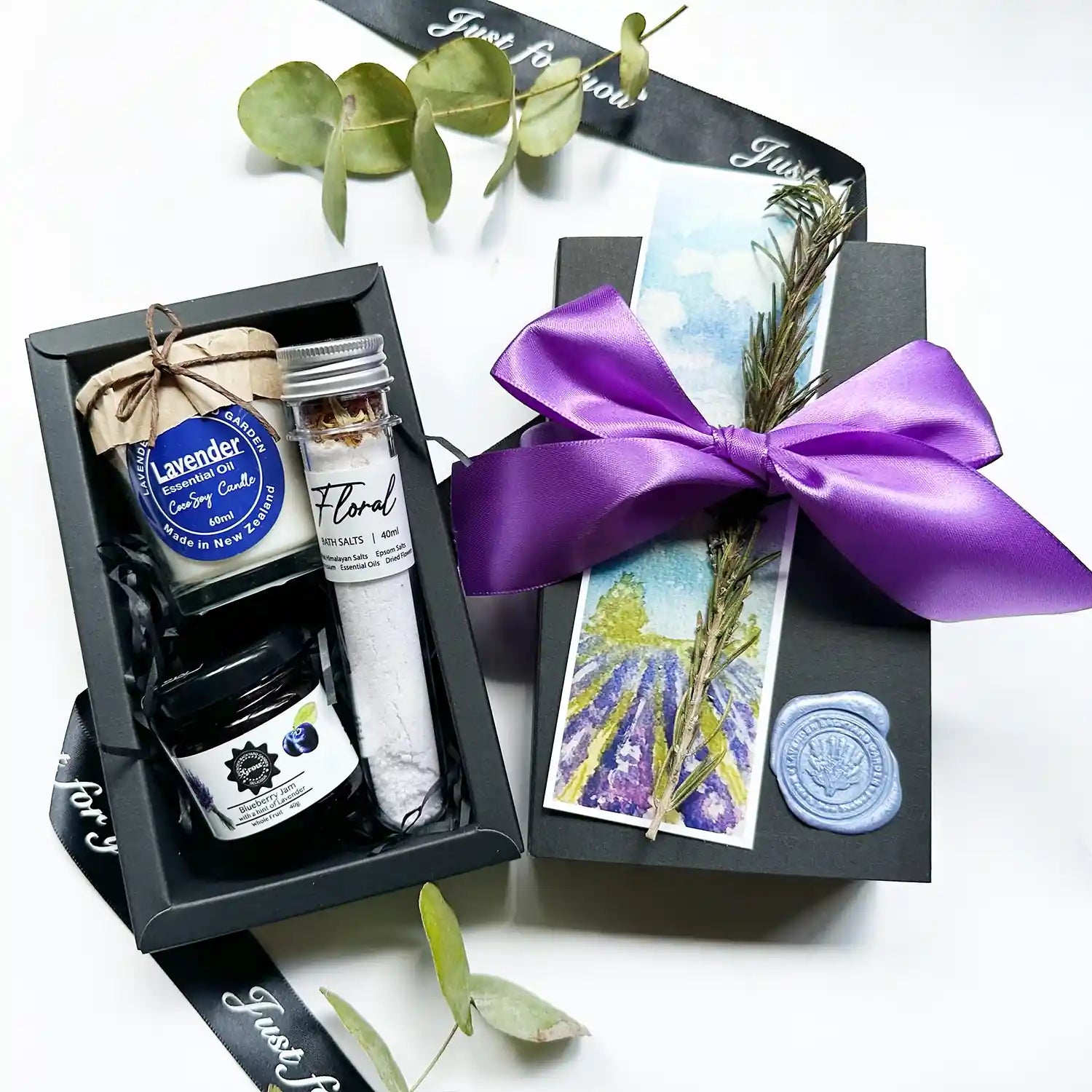 A beautifully wrapped gift box featuring lavender bath salt, lavender blueberry jams, and a soy candle scented with lavender essential oil, elegantly tied with a purple ribbon and complemented by a lavender field bookmark.