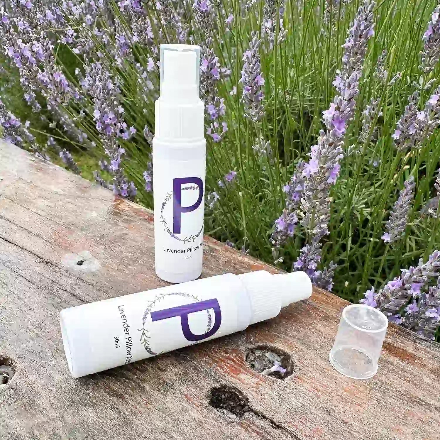 Two bottles of travel-size sleep spray scented with lavender essential oil, placed on a wooden bench with a blooming lavender field in the background at Lavender Backyard Garden, NZ.