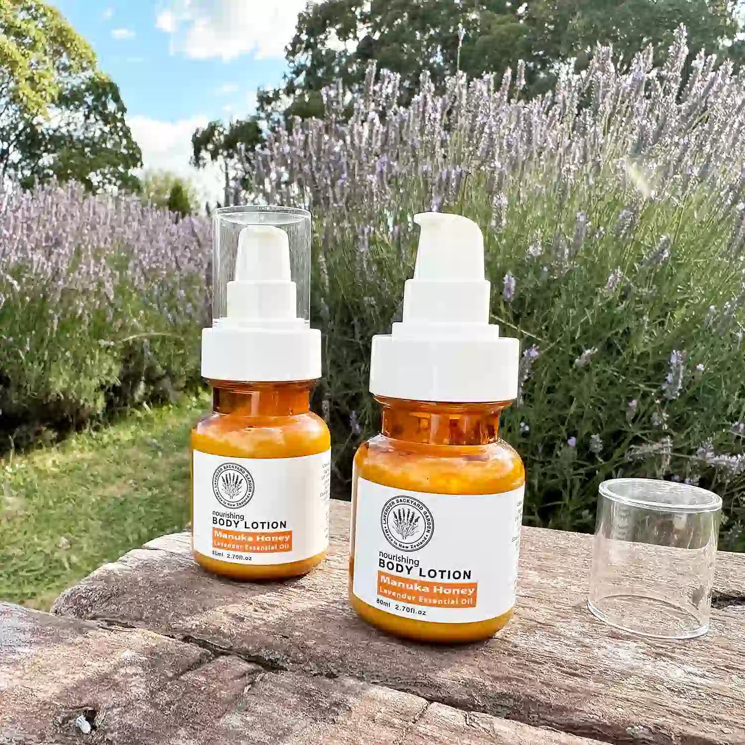 Two bottles of Manuka Honey Lavender Body Lotion, one with lid off, placed on a bench against lavender blooming at Lavender Backyard Garden, New Zealand.