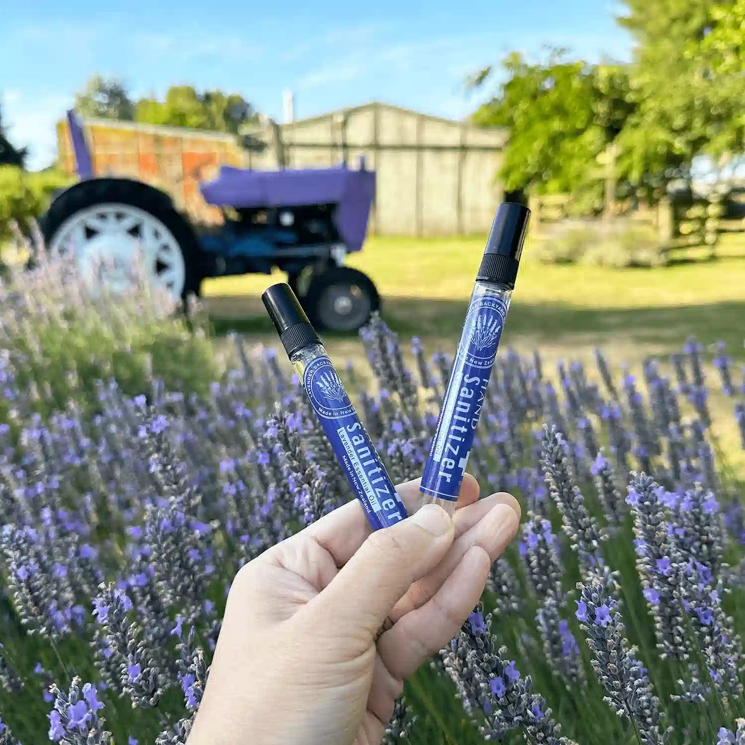  Two Lavender Hand Sanitiser bottles scented by lavender essential oil, held by hand against a backdrop of a lavender field with a farm shed and purple tractor at Lavender Backyard Garden, NZ.