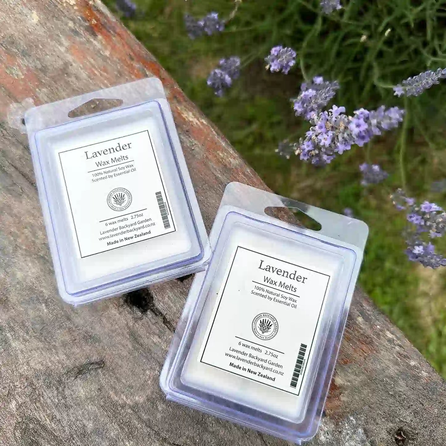 Two packs of lavender wax melts scented with essential oils, placed on a wooden bench with a background of blooming lavender flowers at Lavender Backyard Garden.