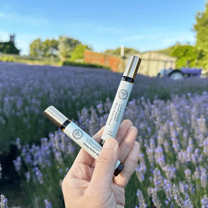 Two Sweet Dream Lavender Sleep Roller Blends, scented by lavender essential oils, held by hand with a background of blooming lavender flowers, a rustic farm shed, and a purple old tractor at Lavender Backyard Garden, NZ.