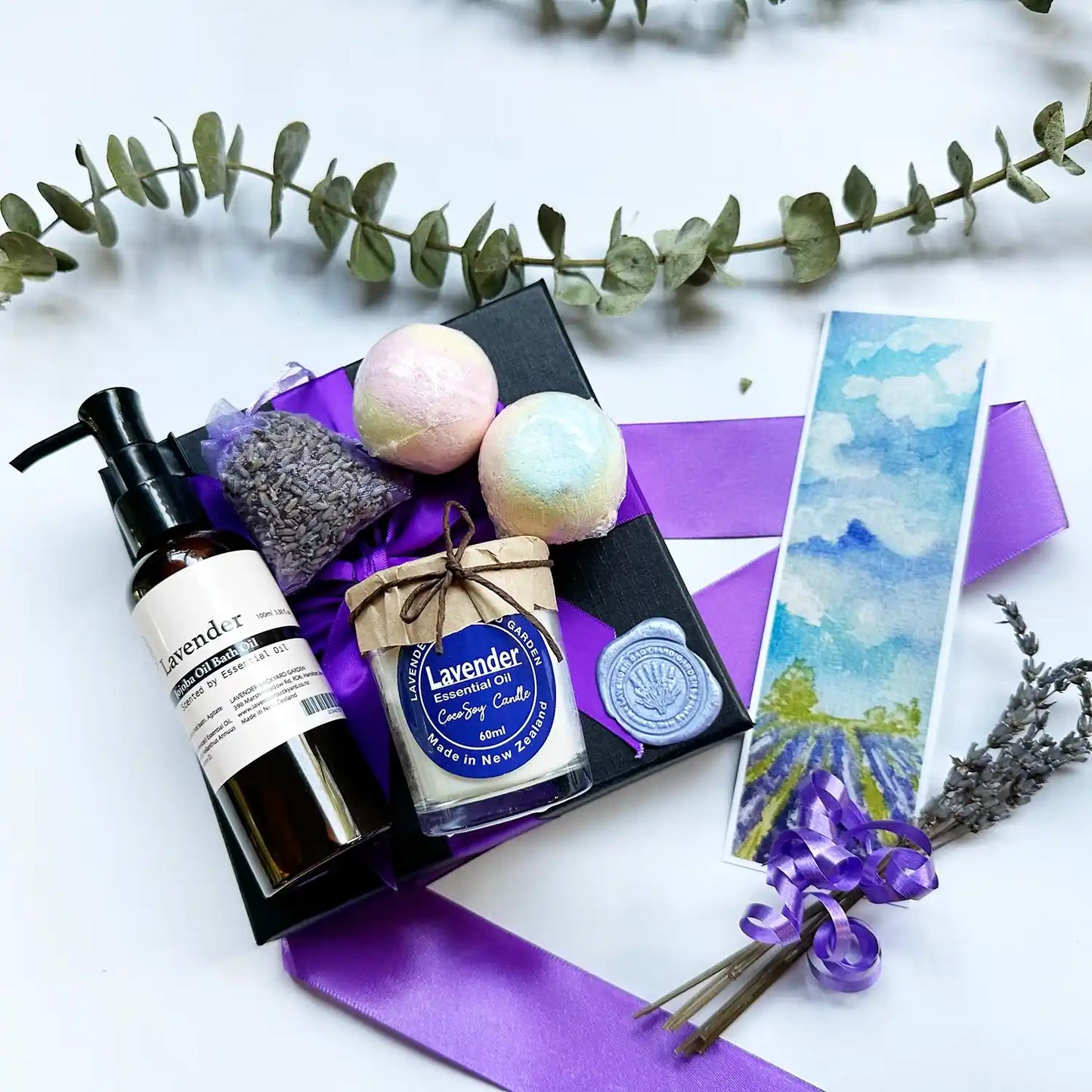 Ultimate Therapy Special Gift for Her featuring a lavender soy candle, bath oil scented with lavender essential oil, bath bombs, and a dried lavender sachet, all beautifully arranged in a purple ribbon-wrapped black gift box.