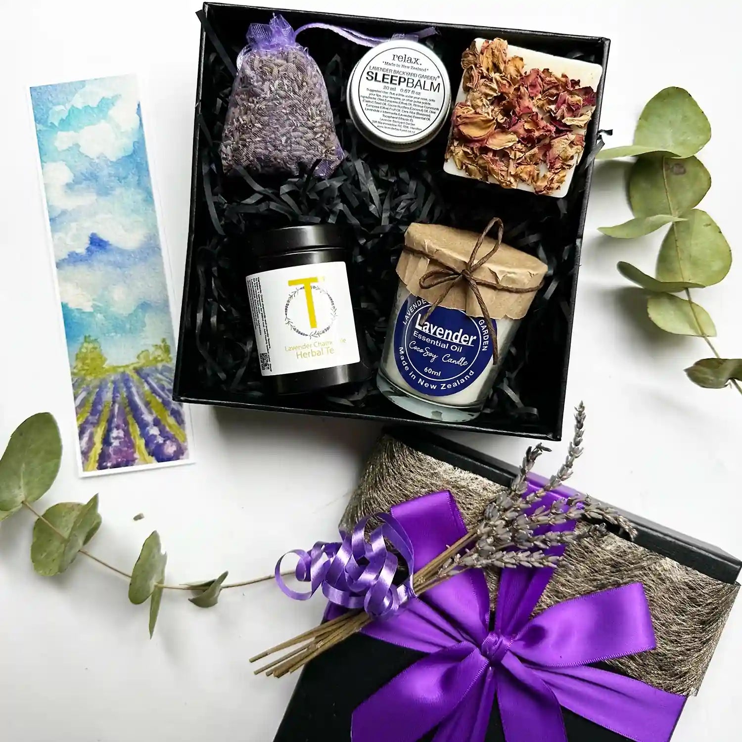 Unique Valentine's Day Gifts box wrapped with a purple ribbon and a black gift box, featuring lavender handmade soap, a lavender soy candle, a soothing sleep balm, lavender chamomile tea, and a lavender sachet, all scented with lavender essential oil, accompanied by a lavender field bookmark.