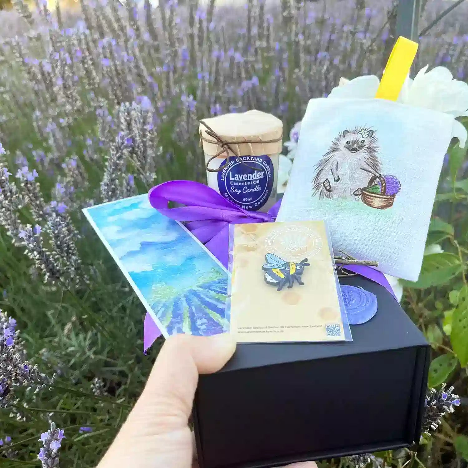 Unwind Lavender Gift Set with Farm Animals placed upon blooming lavender flowers, featuring products such as a Very Cute Bee Badge, Natural Lavender Scented Soy Candles, Lavender Field Bookmark, and Mini Dried Lavender Flower.